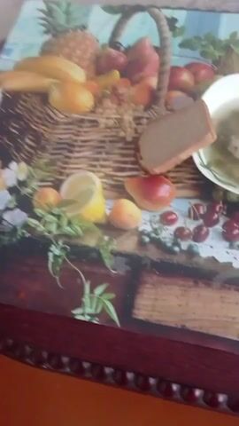 A static overhead shot displays a white bowl of green soup containing meat and vegetables, with a silver spoon resting inside. To the left of the bowl is a slice of dark bread. These items are positioned on a colorful table mat depicting a variety of fruits, including cherries, grapes, apples, and a watermelon slice, alongside a woven basket design. The mat rests on a dark wooden table with an ornate edge. The camera slowly zooms in on the bowl of soup and bread over the duration of the video. No individuals are present.