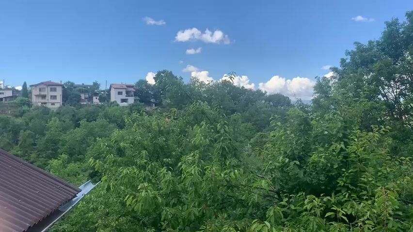 The video shows a lush green landscape with several trees and houses in the background. The sky is blue with scattered white clouds. In the foreground, there is a brown roof and a wooden railing. The middle ground is filled with dense green foliage from the trees. In the background, several houses with white and beige facades are visible on a hill. To the right of the video, a blue car is parked on a dirt road surrounded by grass.