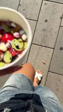 A person's hand, adorned with colorful nail polish and a silver bracelet, holds a white paper cup filled with a frozen dessert. The dessert is topped with an assortment of colorful ingredients, including sliced strawberries, black berries, mini marshmallows, and what appear to be green candies or fruit slices. A bright green spoon is inserted into the dessert. The person is wearing a denim skirt and their legs are visible. The background shows a paved surface, suggesting an outdoor setting. The lighting indicates it is daytime.