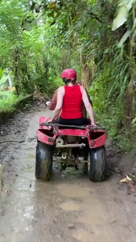 A first-person perspective shows a woman in a red tank top and a red helmet, sitting on a red four-wheeler as it moves through a muddy, dirt path surrounded by a lush green forest. The camera quickly pans upwards, blurring the green canopy of trees overhead. The four-wheeler appears to be driving beside a low, grey concrete wall. The overall impression is of an outdoor adventure through a tropical environment.