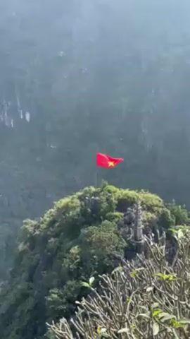 The video shows a panoramic shot of a mountainous landscape, likely taken from an elevated vantage point. In the foreground, there are branches of a small tree, possibly a bush, slightly out of focus.  The focal point is a small mountain peak adorned with lush green vegetation and a weathered stone structure. Atop the peak flies a bright red flag with a prominent yellow star. The flag is the flag of Vietnam. The background consists of more steep, rocky mountains shrouded in a light haze.  The overall impression is one of a serene and somewhat misty landscape.
