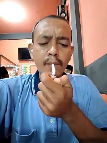 A man with short dark hair and a mustache, wearing a light blue polo shirt, is seen lighting a cigarette and smoking it indoors. He is sitting in what appears to be a small business or store, with a TV visible in the background and shelves stocked with various items. The lighting is bright and overhead. He takes a drag from the cigarette, exhales smoke, and gestures with his hand while talking.