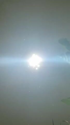 The video shows a close-up, aerial view of sunlight reflecting on the surface of water. The sun is directly in the center of the frame, creating a bright lens flare that extends horizontally across the image. Small particles or bubbles are visible scattered across the water's surface. In the upper right and lower right corners, green leaves, possibly from a lotus plant, are partially visible. The overall impression is of a tranquil outdoor scene, likely a pond or lake, illuminated by the sun.