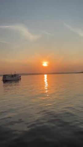 The video shows a serene lake at sunset. The sky transitions from a pale blue to a warm orange as the sun dips towards the horizon. The water is calm, reflecting the colors of the sky. A small white boat is visible in the distance, anchored in the water. Another smaller object that appears to be a floating dock with a bench is also present in the water. There are no people visible in the video. The overall atmosphere is peaceful and tranquil.