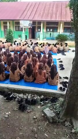 The video shows a large group of students, mostly young people, sitting on blue tarps spread out on the ground in front of a building. The students are wearing similar uniforms, which appear to be brown tops and pants or skirts, and many are also wearing headscarves. They are all facing the building. Shoes are neatly arranged in rows on the ground in front of the tarps. The building is a single-story structure with a red roof and green and white walls. There are windows and a doorway visible. Posters are displayed on the wall of the building. The ground is a mix of dirt, gravel, and some vegetation. The overall impression is of an outdoor school gathering or assembly.