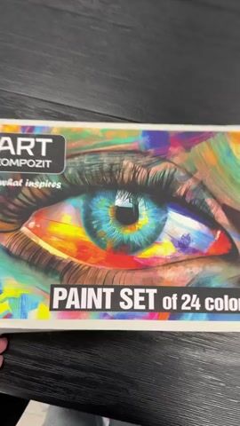 The video begins with a close-up of a box for an ART KOMPOZIT paint set of 24 colors. The box features an abstract eye design. A hand opens the box, revealing rows of small paint pots. The camera then pans across the open box, showing the lids of the paint pots, each labeled with a number and color name. Some of the visible colors include pink, yellow, deep green, carmine, and black. The paint pots are arranged in a grid within the cardboard box. The background is a dark, textured surface, likely a table.