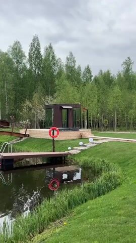 The video shows an outdoor scene on a cloudy day. A pond reflects the sky and surrounding greenery. There is a small wooden bridge crossing the pond, leading to a small island with a fountain. On the pond's edge, there is a wooden dock with a ladder leading into the water. A life preserver is mounted on a post nearby. Adjacent to the pond, there is a small, modern cabin-like structure with glass walls and a dark exterior. Tall green trees are visible in the background, surrounding the area. The grass is well-maintained, and there are decorative white spheres scattered around the cabin. The camera pans slightly to show the overall landscape.