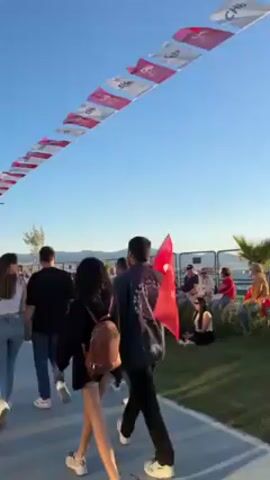 Here is a concise description of the video:

The video captures a scene on a sunny day, likely at an outdoor event or celebration. The sky is clear and blue, providing a vibrant backdrop to the scene. A row of white and red flags is strung overhead, creating a festive atmosphere. A group of people, including couples and individuals, are walking along a paved path. One person is carrying a Turkish flag. To the right of the path, people are seated on chairs on a patch of grass. They are likely spectators or participants in the event. A fence runs along the back of the grass area, separating the event from the water and hills in the distance. The overall atmosphere is one of community and celebration.