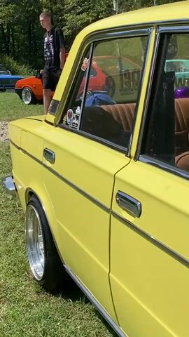The video shows a close-up of the side of a yellow vintage car. The car is parked on grass, and other vintage cars are visible in the background. The car's interior is visible through the windows, including brown fabric seats. A yellow keychain in the shape of a cartoon character hangs from the rearview mirror. Two men are partially visible in the background, one wearing a white t-shirt and the other a red t-shirt. The scene appears to be an outdoor car show or gathering.