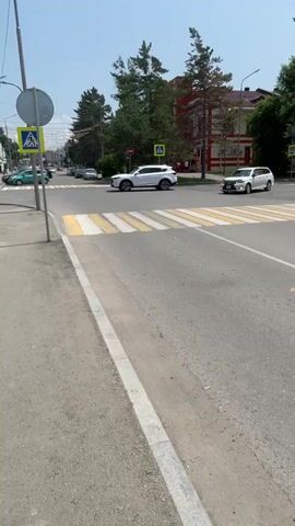 The video shows a street scene on a sunny day with trees and buildings in the background. There are two cars visible, a white SUV and a white minivan, driving on the road. A pedestrian crossing is marked by yellow and white stripes. In the distance, there are several pedestrian crossing signs. The camera pans across the scene, showing the cars and the road.