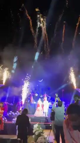This video shows a wedding celebration. In the center is a couple standing on a platform, illuminated by blue stage lights. Pyrotechnics and small vertical fountains of sparks frame them and shoot up into the air. Surrounding the platform, people are taking pictures and recording with their phones. The background is dark, with silhouettes of structures and performers. In the foreground, individuals are seated, partially visible, observing the event.