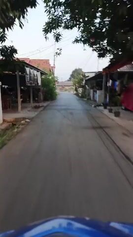 This is a first-person perspective video of a person on a scooter or bicycle moving down a paved road. The road is lined with single-story buildings on both sides. Some of the buildings appear to be shops or small businesses, with awnings and signs visible. There are trees and vegetation growing along the sides of the road and near the buildings. Overhead, there are numerous electrical wires and poles. The sky is overcast or hazy, suggesting early morning or late afternoon. In the distance, the road appears to curve slightly and lead towards a slightly more open area with additional buildings and structures. There are no other vehicles or people clearly visible in the immediate vicinity. The overall environment suggests a residential or mixed-use area in a village or small town.