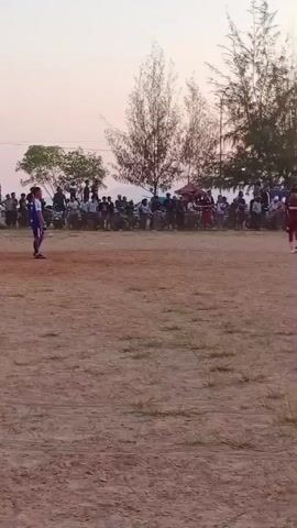 A man in a red jersey is running across a dirt field while a crowd watches from the sidelines. The man is in the middle of the field, running towards the right of the frame. Behind him, a crowd of people are gathered, some sitting and some standing. They appear to be watching the man play a sport. The background consists of trees and a building with a light blue roof. The sky is a pale orange, suggesting it is either sunrise or sunset. The field is mostly dirt with patches of dry grass. There are a few discarded plastic bottles scattered on the ground.