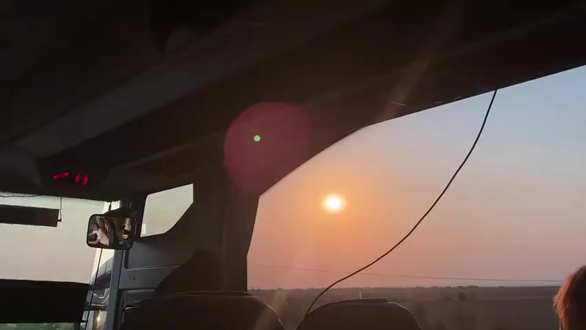 The video is shot from inside a vehicle, likely a bus or a van, looking out a window. The sun is visible in the sky, appearing as a bright, circular orb, and it is setting or rising, casting a warm, orange glow across the horizon. The sky transitions from a clear blue at the top to a gradient of orange and pink closer to the horizon. In the foreground, the interior of the vehicle is visible, including seats, a dashboard with lights, and a side mirror reflecting a person's face. A black cable hangs down from the ceiling of the vehicle, partially obscuring the view of the sunset. The landscape outside appears to be rural, with some trees and what might be fields or open land. There is a lens flare, appearing as a pinkish circle with a green dot in the center, on the window. The overall impression is one of travel during a scenic sunset or sunrise.