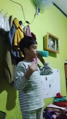 A young boy with dark hair is standing in a room with lime green walls. He is wearing a grey and white striped long-sleeved shirt and matching pants. He is holding a cylindrical object, possibly a spray can or bottle, in his right hand and is moving it in a circular motion above his head. His left hand is resting on his chest. In the background, there are several items of clothing hanging on a rack on the wall. To the right of the boy, there is a whiteboard with some markings on it and a pile of clothes on the floor. A framed picture depicting a mosque is hanging on the wall above the whiteboard. A ceiling fan is visible in the upper right corner of the frame. The boy appears to be counting or reciting something as he performs the action.