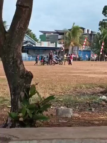 The video shows an outdoor scene, possibly a field or park, with a group of people and several motorcycles. Indonesian flags are displayed along a fence or barrier in the background, suggesting a festive or patriotic occasion. The ground is covered with dry grass and some debris. A large tree is visible in the foreground, partially obscuring the view. Some people are standing next to motorcycles, while others appear to be riding them or moving around the area. The sky is overcast, and the overall atmosphere seems casual and social.