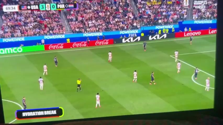 A soccer match is being shown on a screen, with the score United States 3, Paraguay 0. The game is in its 69th minute, and there is a hydration break. Several players in white and blue uniforms are on the green field, along with a referee in a yellow shirt. A large crowd is visible in the stadium seats behind the field. Advertising banners are displayed around the stadium.