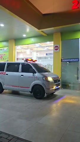 The video captures an ambulance parked in front of a hospital entrance at night. The ambulance is white with red and grey stripes and the word "AMBULANCE" printed on its side. It has flashing red and blue lights on its roof and headlights. The hospital entrance features large glass doors and windows, with signs indicating "IGD 24 JAM" (24-hour emergency department) and "DEKONTAMINASI" (decontamination). The overall setting appears to be a well-lit medical facility. The time of day is clearly nighttime due to the artificial lighting and the dark exterior. No people are visible in the frame. The atmosphere is that of a functional, operational hospital, likely prepared for emergencies. The location cues suggest Indonesia.