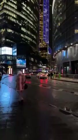 A rainy night scene in a modern city is captured in a vertical video. The street is wet, reflecting the vibrant city lights. Tall skyscrapers with illuminated windows loom on both sides, creating a canyon-like effect. Traffic moves along the wet road, including taxis. People are walking on the sidewalks, some carrying umbrellas. One person in a pink and white outfit stands in the foreground, looking towards the street and skyscrapers. The overall atmosphere is dark, with the city lights providing a strong contrast.
