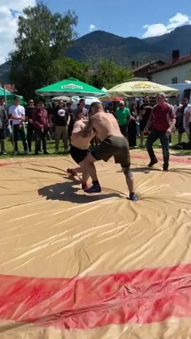 Two shirtless men are wrestling in the center of a beige and red wrestling mat, surrounded by a crowd of spectators. The man on the left wears black shorts, and the man on the right wears dark green shorts and blue wrestling shoes. The man on the right has tattoos on his arms and back. They grapple and circle each other on the mat. The crowd of spectators surrounds the mat, some taking photos or videos. In the background, there are several green and white tents, houses, and green mountains under a blue sky. The environment appears to be outdoors, possibly a fair or festival.