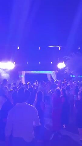 The video shows an outdoor nighttime gathering with many people dancing. The scene is illuminated by a stage with yellow fabric drapery and a backdrop, featuring bright purple and green lights and spotlights. String lights are visible overhead. The people are dressed in casual attire, with some wearing hats and dresses. The ground appears to be paved with stones. The atmosphere suggests a party or celebration, possibly a wedding reception or a similar event. The camera is positioned behind a group of dancers, looking towards the stage.