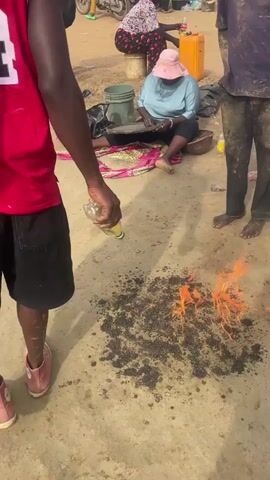 The video shows a group of people outdoors. A small fire burns on the ground as a man wearing a red basketball jersey and black shorts pours liquid from a plastic bottle onto the flames. Another man stands barefoot nearby, covered in dirt. A woman sits on a blanket, appearing to sift through something on a pan. Other people are visible in the background, along with a motorcycle and various containers. The ground is a mixture of dirt and concrete.