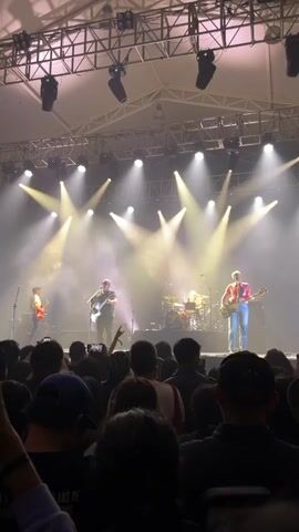 A full shot captures a live band performance on a brightly lit stage, seen from the audience's perspective. The stage is bathed in white spotlights and filled with smoke. Five band members are visible: two guitarists, a bassist, a drummer, and another musician who appears to be playing a guitar, though they are further back on stage. The audience, comprised of numerous people, stands close to the stage, many holding up their phones to record the performance. The architectural details of the venue, including the lighting rigs and structural beams, are visible above the stage.