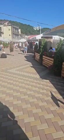 The video shows a paved walkway lined with businesses and trees. People are walking along the path, some sitting at tables outside a cafe, and a child is riding in a small toy car. The area appears to be a resort or tourist destination with buildings and lush greenery in the background. The sky is clear and blue, indicating a sunny day.