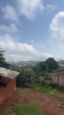 This media file captures a view of a village nestled on a hillside under a partly cloudy sky. The scene is dominated by a sprawling collection of buildings with predominantly reddish-brown brick walls and various roof types, including corrugated metal and tiled structures. Lush green vegetation, including banana plants and trees, is interspersed throughout the village and in the foreground.

In the immediate foreground, a dirt path leads uphill. To the left, a small, rustic building with a corrugated metal roof is visible. To the right, a neatly constructed brick wall marks a boundary. The overall impression is one of a densely populated residential area integrated with its natural surroundings.

The time of day appears to be daytime, with bright sunlight casting shadows, suggesting it's likely morning or afternoon. The sky is a vibrant blue, punctuated by fluffy white clouds, indicating fair weather. The location cues—the architectural style, the prevalent vegetation, and the hilly terrain—point towards a tropical or subtropical region. Gembu, Nigeria, is provided as the location context.

No people are visible in the media file, and no specific activity or event is clearly taking place. The scene is static and observational. The emotions conveyed are neutral, presenting a straightforward depiction of the village landscape. There is no visible text within the media file.