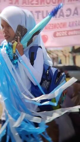 A young woman wearing a white hijab and white long-sleeved shirt, and dark pants, walks while talking on her phone. She has a backpack on her back and is holding a blue and teal umbrella decorated with blue streamers and blue beaded accents in her right hand. She is also holding a yellow lanyard with a charm. The background is blurred with hints of trees and a banner with text and cartoon characters, suggesting an outdoor event or parade. The lighting is bright, indicating daytime.