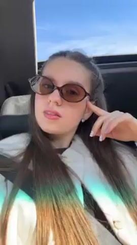 A young woman wearing sunglasses and a white jacket sits in the driver's seat of a car and faces the camera. The car's sunroof is open, and the sky is visible.  In the backseat, a toddler is secured in a car seat. The woman smiles and poses slightly for the camera.  Music is playing in the background. The sunlight is streaming in and creating lens flare effects.