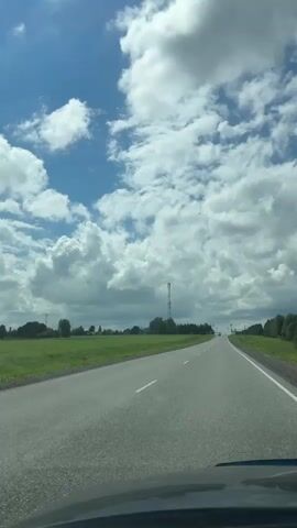 The video captures a view from a moving vehicle on a paved road. The sky is predominantly blue with scattered white, fluffy clouds. On either side of the road, there are fields of green grass. In the distance, there is a tall, slender tower, possibly a communication tower, and a few trees. There are other vehicles visible on the road ahead. The overall impression is of a rural landscape on a partly cloudy day.