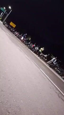 Here is a description of the video.

The video shows a night scene on a paved road. A row of parked motorcycles lines the edge of the road, next to a white line. Several people are standing or walking among the motorcycles. One person is wearing a bright yellow shirt or poncho. The background is dark, suggesting it is nighttime, and there are streetlights illuminating the area. There is also a road sign in the distance. The video camera is handheld and is shaking.