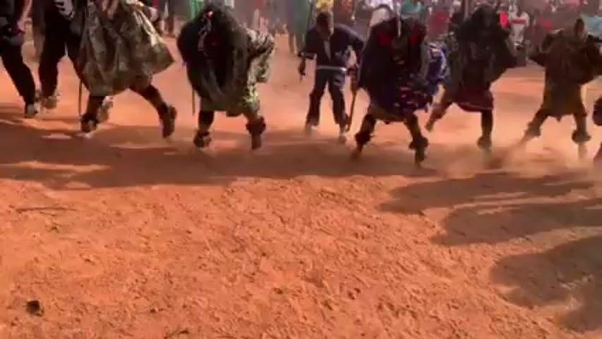 The video depicts a cultural performance or ceremony taking place in an open, dusty area with a crowd of people observing in the background. Several individuals, some wearing elaborate masks and costumes, are dancing or moving rhythmically, kicking up dust. Some carry sticks. The backdrop includes simple structures and a large gathering of spectators. The overall impression is of a vibrant, possibly traditional, outdoor event.