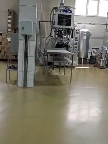 The video pans across a clean, brightly lit industrial interior, likely a food processing or packaging facility. The floor is a smooth, pale beige epoxy, reflecting light. Dominating the scene are several large, stainless steel machines, appearing to be packaging or filling equipment. One prominent machine features a vertical column, a control panel with buttons and a screen, and a mechanism at the top that seems to be dispensing or processing a substance. Next to it, a tall, cylindrical stainless steel tank is visible. In the background, stacks of cardboard boxes are arranged on shelves. There are no people visible in the video. The environment is sterile and organized, suggesting efficient operation. The lighting appears to be from overhead fluorescent fixtures and natural light from windows.