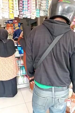A person wearing a black hoodie and jeans, with a black helmet on, stands facing away from the camera, holding a plastic-wrapped item in their right hand. The person is in a store, which appears to be a convenience store or a small grocery store. Shelves are visible behind the person, stocked with various packaged goods. To the left of the frame, a person wearing a hijab and a patterned top is interacting with another person who is also partially visible. The floor appears to be tiled. The lighting suggests it is daytime.