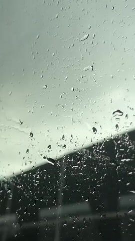 The video shows a close-up of raindrops on a window. The sky is overcast and gray, with the light reflecting off the wet glass. The raindrops are of various sizes, some are large and clustered together, while others are small and scattered. The background is blurred, but appears to be an outdoor scene with dark shapes that could be buildings or trees. The overall impression is one of a rainy day.
