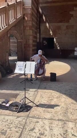 The video shows a man playing the cello in the courtyard of the Palazzo della Ragione in Verona, Italy. He is seated in the center of the frame, dressed in white with a white cap, and playing his cello. A music stand with sheet music sits in front of him, displaying a QR code for donations. A donation box is on the ground. In the background, a staircase leads up to the Palazzo, and another man is climbing the stairs. The camera then pans up to show the exterior of the Palazzo della Ragione against a clear blue sky. The building has a distinctive striped pattern and arched windows.
