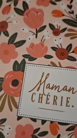 The video is a close-up, slightly shaky shot of a floral-patterned notebook or card. The background features a pink base with scattered illustrations of coral and orange flowers with dark green leaves. In the center of the frame, a white rectangular label with a dotted border is affixed. The label contains two lines of text written in a golden, cursive font. The top line reads "Maman" and the bottom line reads "CHÉRIE." followed by an underline. The camera remains static for the duration of the clip.