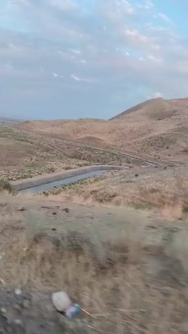 A drone shot pans across a dry, rolling landscape. The ground is covered in sparse, golden grass, with a few scattered green shrubs. A small, blue body of water is visible in the middle ground, bordered by more dry vegetation. In the background, gentle hills rise towards a pale sky. The scene appears tranquil and untouched by human activity, with no visible structures or people.