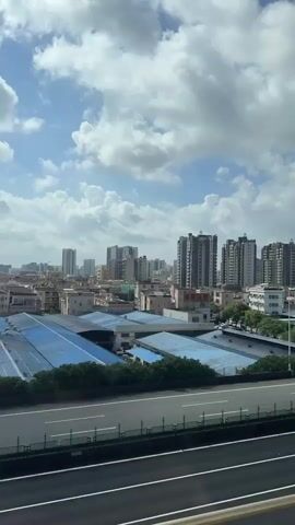 The video shows a cityscape with a highway in the foreground. Several tall buildings are visible in the distance, along with smaller buildings and trees. There are clouds in the sky. Cars and a truck are driving on the highway.