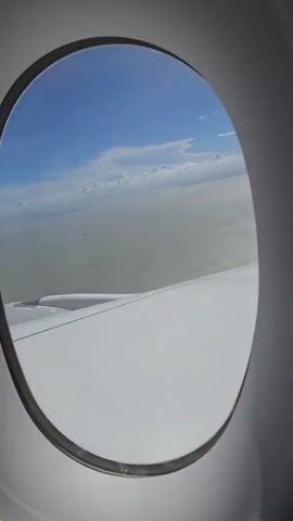 The video shows a view from an airplane window looking out at the ocean and sky. The wing of the airplane is visible in the foreground. The sky is blue with some white clouds. The ocean is a hazy green color. There appear to be some ships in the distance on the water. The video is stable and does not show any significant movement.