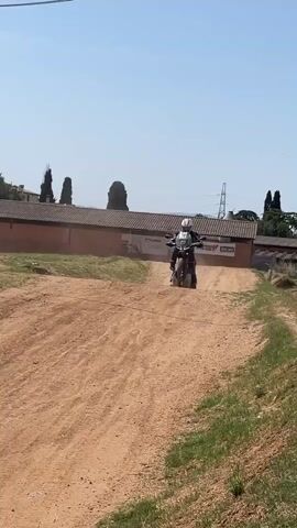 The video shows a motorcycle jumping over a dirt hill. The rider is wearing a white helmet and protective gear. The motorcycle has the number 51 on its side. After the jump, the rider lands and continues down the dirt path. The camera then pans to show two people sitting on the ground, watching the motorcycle. One person has a camera. The background includes trees, a building with a tiled roof, and power lines. The sky is clear and blue.