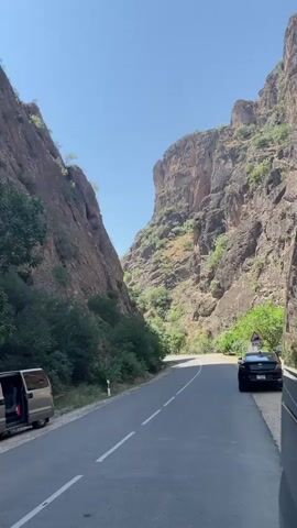 The video shows a narrow road with steep, rocky cliffs on either side. A silver van with its side door open is parked on the left side of the road. A black car is parked on the right side of the road. The sky is clear and blue. The sun is shining, casting shadows on the cliffs. There is vegetation growing on the cliffs and along the sides of the road. The road appears to be winding through a canyon. The camera is looking forward down the road.