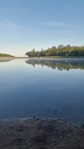 A calm lake reflects a treeline and a clear blue sky. In the foreground, there is dark, rocky soil. A faint mist hovers over the water's surface. The video is of a natural landscape at sunrise or sunset, with no people or animals visible.
