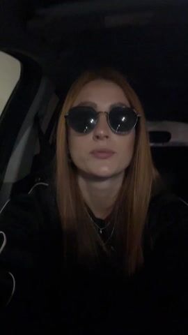 A red-haired woman wearing sunglasses and a black track jacket is inside a car. She is singing and holding a phone to record herself. The car’s interior is mostly black. The camera is handheld and shaky.