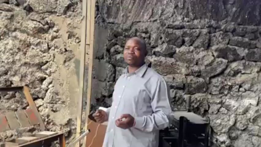 A man is standing outdoors in front of a stone wall. He is wearing a light gray long-sleeved shirt. To his left is a metal contraption with wood. To his right is a metal structure with a dark object. The man is gesturing with his hands as he speaks. The background includes two different stone walls.
