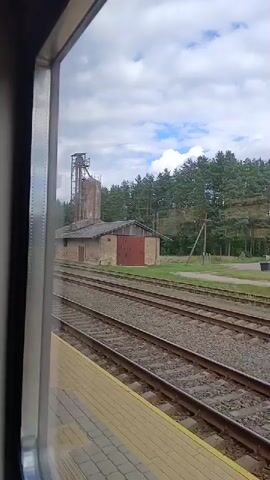 The video shows a view from a train window, looking out at a rural landscape. In the foreground, there are train tracks and a yellow platform. Beyond the tracks, there is a building with a red door and a tall, slender structure next to it. Behind the building, there is a dense forest of green trees, and the sky is filled with white clouds. The camera is moving, indicating the train is in motion.