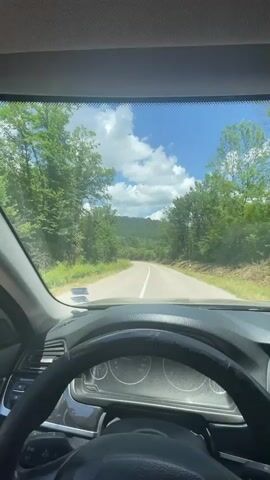 The video shows a first-person perspective of driving a car on a paved road through a wooded area. The sun is shining, and there are white clouds in the blue sky. The road curves ahead, leading into a hilly, forested landscape. The dashboard and steering wheel of the car are visible in the foreground.