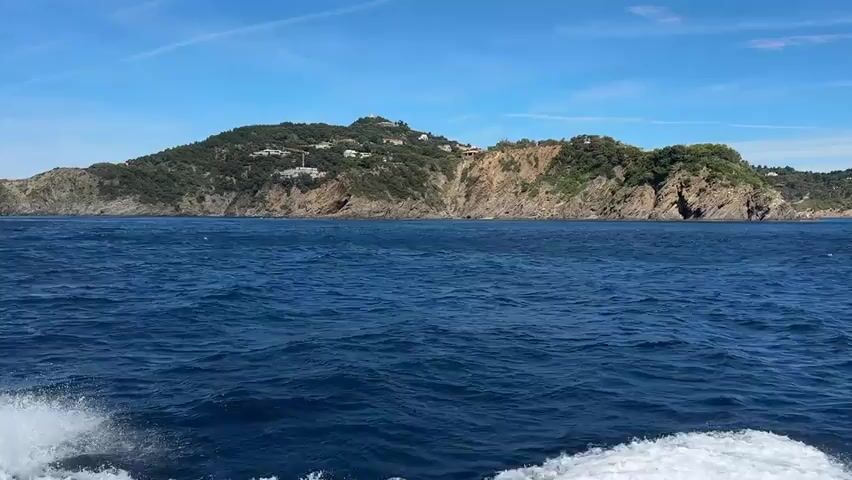 A wide-angle, first-person perspective video shows a boat moving through the choppy blue ocean. The wake of the boat is visible in the lower left corner of the frame. In the distance, a green, hilly island with rocky cliffs and scattered buildings rises from the sea under a clear blue sky with some wispy clouds. The island is covered with trees and vegetation, and some structures are visible on its slopes. The sun is shining, casting a bright light on the scene. The camera is pointed towards the island, capturing the expanse of the sea and the approaching landmass.